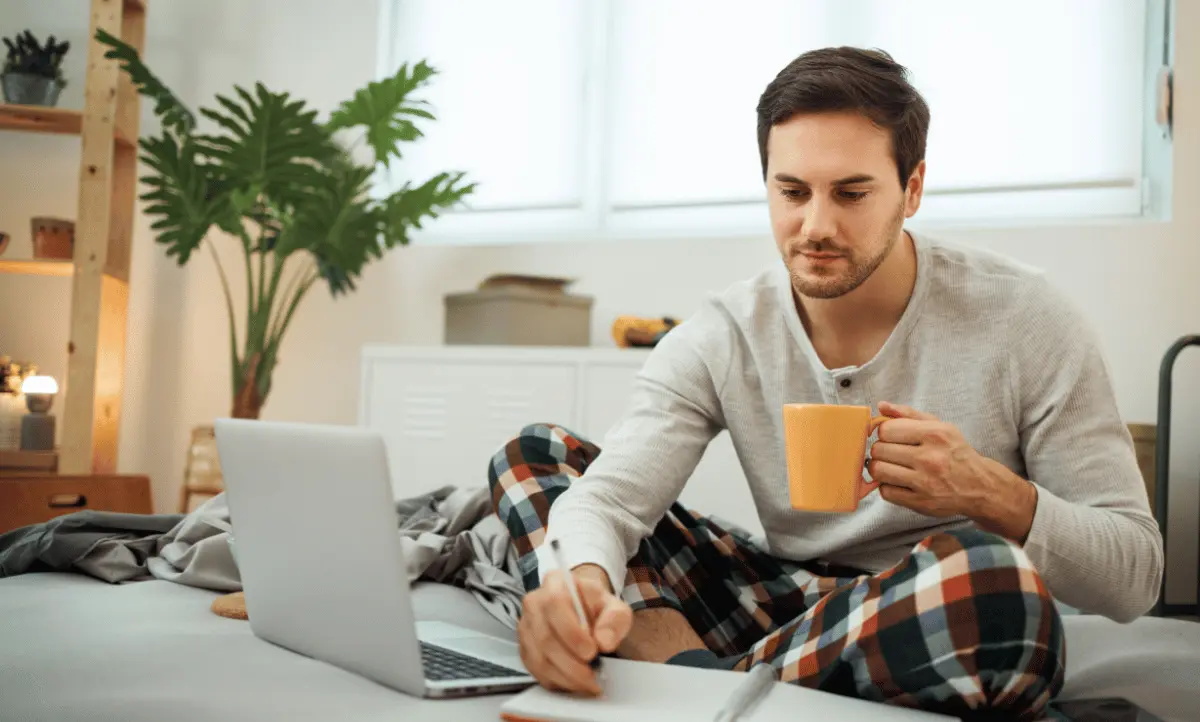 Een man zit ontspannen op bed in comfortabele kleding, met een kop koffie in de hand en schrijft in een notitieboek, terwijl een laptop voor hem openstaat. Ideaal voor een rustige en productieve ochtendroutine.