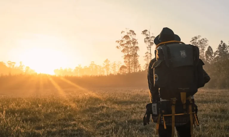 Een solo-reiziger met een grote rugzak staat in een open veld bij zonsopgang, genietend van het uitzicht en de rust.