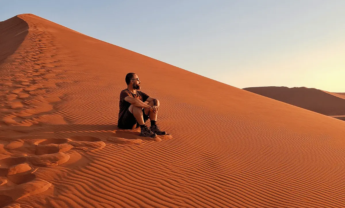 Een man zit ontspannen op een zandduin in de woestijn tijdens zonsondergang, genietend van de rust en het uitzicht, symbool voor solo reizen en zelfreflectie.