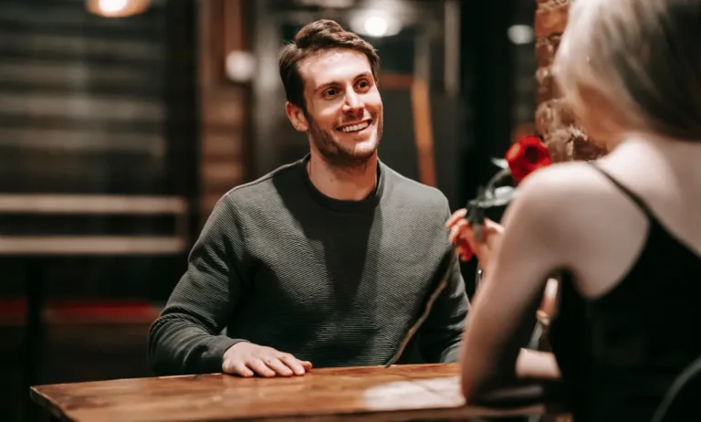 Een man glimlacht tijdens een eerste date in een sfeervolle setting, gekleed in een nette trui, terwijl hij tegenover zijn date zit.