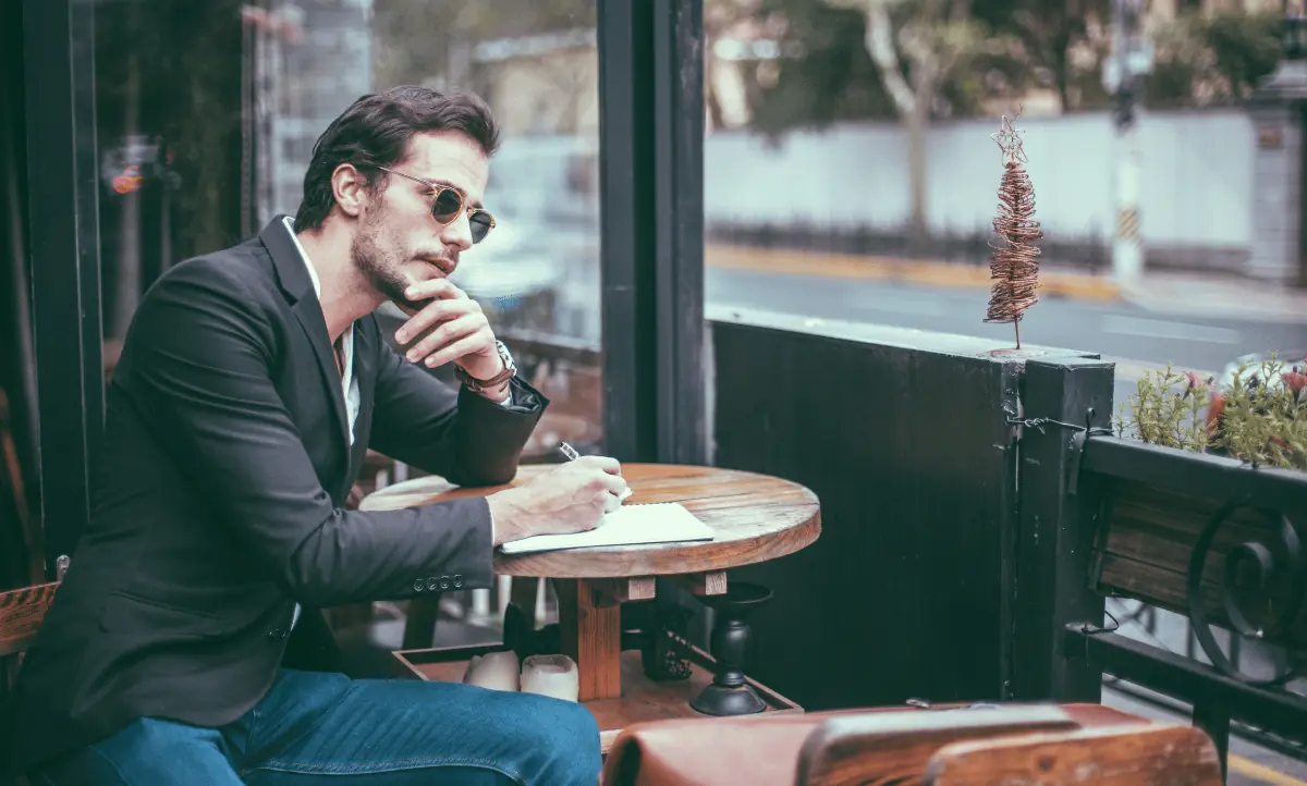 Man schrijft in zijn notitieboekje terwijl hij aan een tafel zit, met een stadse achtergrond, voorbereidend op een solo reis.